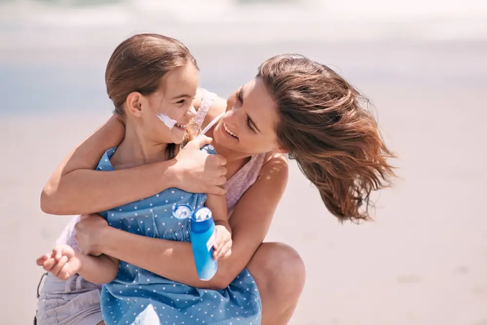 Matka s dcerou na pláži mazající se opalovacím krémem.