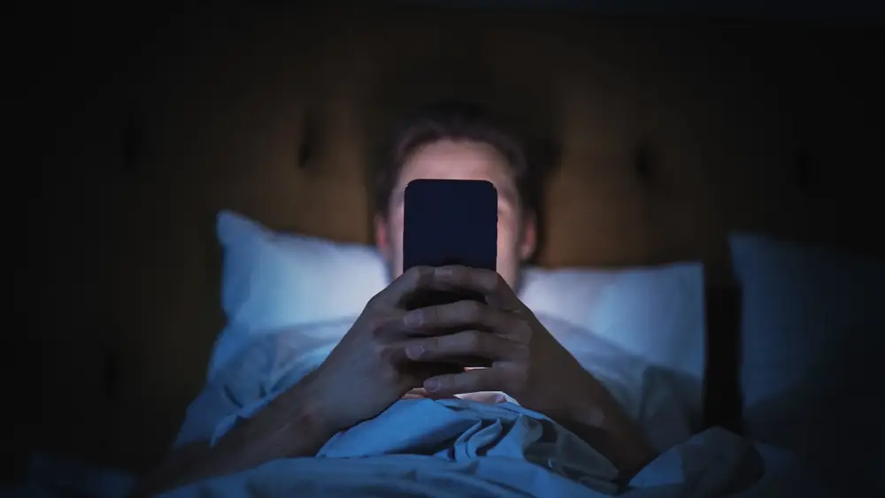 muž ležící v posteli s mobilem v ruce
